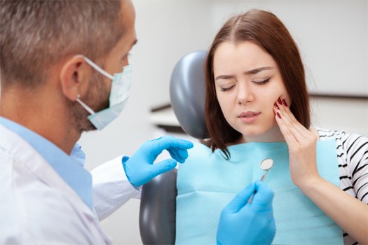 Woman in dental chair with eyes closed pressing hand to cheek to show dentist where it hurts